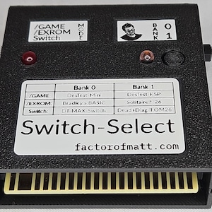 Pode incluir: Um cartucho eletrónico retangular preto rotulado como Switch-Select com uma borda de conector de cor dourada. O dispositivo possui duas luzes indicadoras vermelhas, um botão de pressão e uma etiqueta branca detalhando as configurações de Bank 0 e Bank 1 para os modos /GAME, /EXROM e Switch, incluindo o texto factorofmatt.com.
