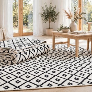 Pode incluir: Um tapete retangular com um padr&atilde;o repetitivo de diamantes pretos sobre um fundo branco. O tapete &eacute; feito de um material t&ecirc;xtil tecido e &eacute; exibido tanto estendido no ch&atilde;o quanto dobrado sobre si mesmo para mostrar a espessura e textura.