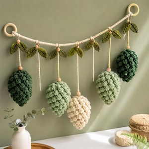 Op de afbeelding: Een wandhanger bestaande uit vijf gehaakte dennenappels in de kleuren donkergroen, lichtgroen en crème. Elke dennenappel hangt aan een gevlochten koord met houten kralen en gehaakte groene blaadjes, bevestigd tussen twee houten ringen.