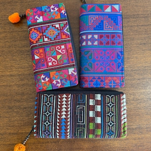 May include: Three rectangular zippered wallets featuring intricate woven geometric patterns. One wallet displays vibrant red, orange, and blue squares. Another features blue, red, and green motifs. The third has a dark base with brown, green, and white stripes.