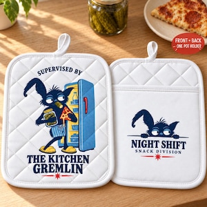 May include: Two white quilted pot holders featuring a blue graphic of a furry creature. One shows the creature holding a jar of pickles and a slice of pizza by an open fridge with the text SUPERVISED BY THE KITCHEN GREMLIN. The other shows the creature peeking over a line with the text NIGHT SHIFT SNACK DIVISION.
