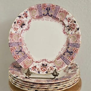 May include: A stack of six round ceramic plates with scalloped edges. The plates feature a cream-colored center and a wide border decorated with pink, purple, and dark blue floral patterns, gold scrollwork, and intricate filigree details.