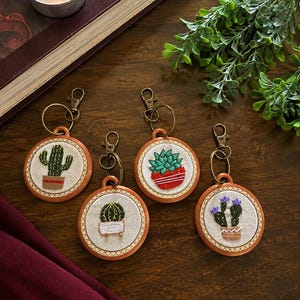 Puede incluir: Cuatro llaveros circulares con diseños de cactus bordados a mano sobre tela blanca, montados en pequeños bastidores de madera. Cada llavero incluye un mosquetón y una anilla de metal color bronce, mostrando varios cactus verdes en macetas con detalles en rojo, blanco y marrón.