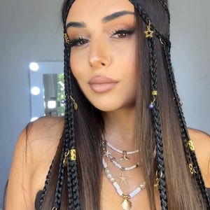 Può includere: Un set di accessori per capelli con molteplici ciocche intrecciate nere decorate con ciondoli in metallo dorato, tra cui stelle, piccole perline a forma di occhio blu e un ciondolo centrale a cuore rosso sulla fronte, con collane di perle bianche.