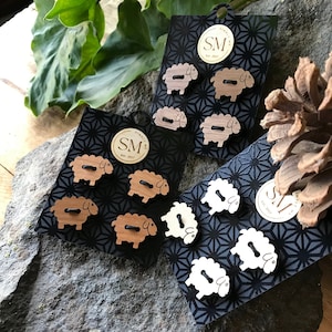 May include: Set of six wooden sheep buttons, three brown and three white, with black stitching for eyes and mouths. The buttons are displayed on black cards with a white geometric pattern and the text "SM* Est. 2017".