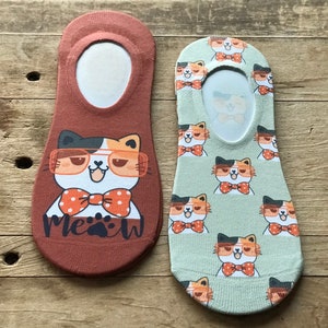 Puede incluir: Dos pares de calcetines invisibles con diseños de gatos. Un par es marrón con un solo gato que lleva gafas y una pajarita con la palabra "Meow" impresa debajo. El otro par es verde con un patrón repetido de gatos que llevan gafas y pajaritas.