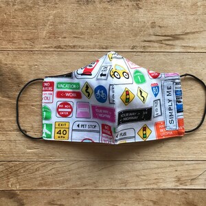 May include: A white face mask with a colorful pattern of road signs, including "Vacation", "Your Way", "Highway", "Exit", "Pet Stop", "Fuel", and "Simply Me".