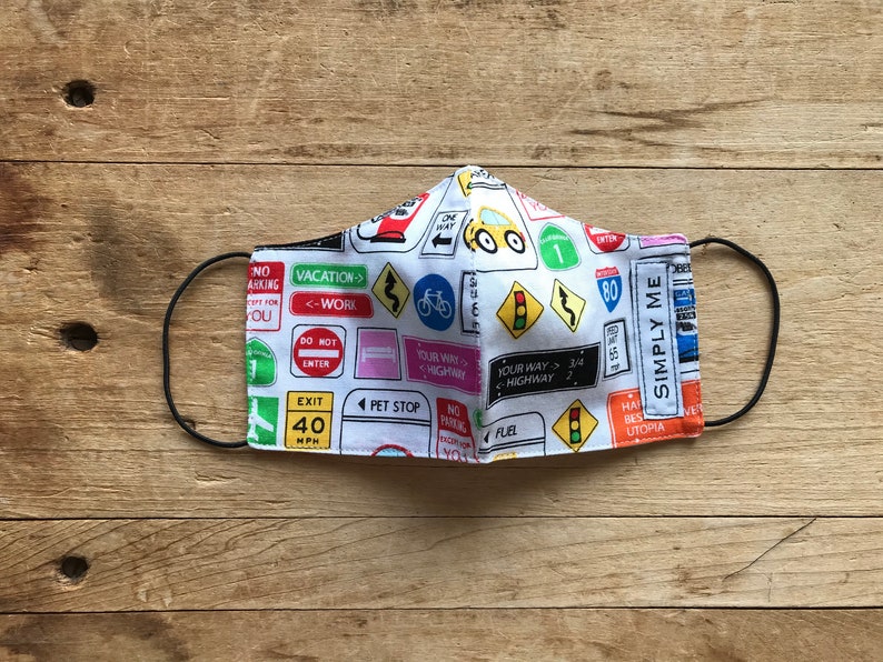 May include: A white face mask with a colorful pattern of road signs, including "Vacation", "Your Way", "Highway", "Exit", "Pet Stop", "Fuel", and "Simply Me".