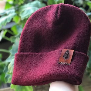 May include: A burgundy beanie with a brown leather patch featuring a tree and the text "Live Simply".