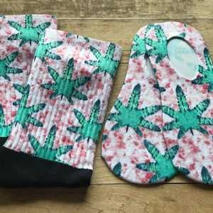 May include: A pair of white socks with a green and red tie-dye pattern featuring a marijuana leaf design. The socks are shown on a wooden surface.