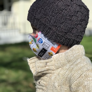 May include: A child wearing a black knit beanie and a white sweater with a face mask featuring a colorful design of traffic signs and the text "SIMPLY ME" and "HA BES UTOPIA".
