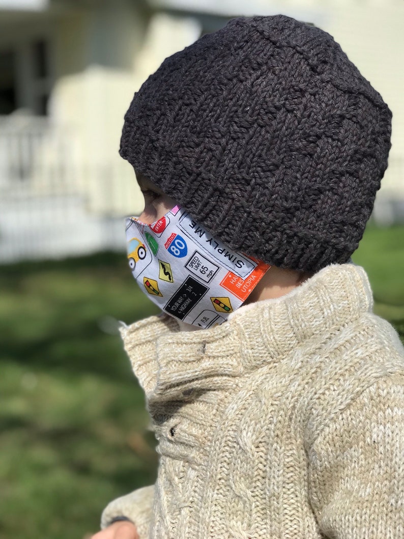 May include: A child wearing a black knit beanie and a white sweater with a face mask featuring a colorful design of traffic signs and the text "SIMPLY ME" and "HA BES UTOPIA".