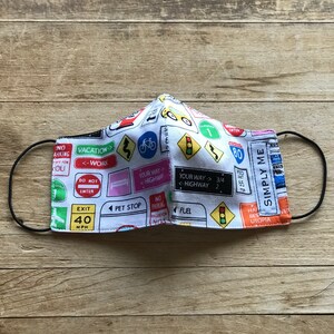 May include: A white face mask with a colorful road sign pattern. The signs include "Vacation", "Work", "Your Way", "Highway", "Exit 40 MPH", "Pet Stop", "Fuel", "Simply Me", and "80".