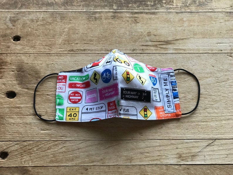 May include: A white face mask with a colorful road sign pattern. The signs include "Vacation", "Work", "Your Way", "Highway", "Exit 40 MPH", "Pet Stop", "Fuel", "Simply Me", and "80".