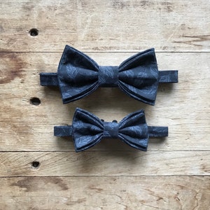 May include: Two black bow ties with a leaf pattern. The larger bow tie is on top of the smaller bow tie.