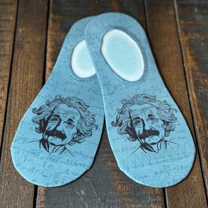 May include: A pair of blue no-show socks with a graphic print of Albert Einstein's face. The socks have a white background with black outlines and a pattern of mathematical equations.