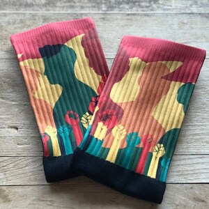 May include: A pair of red, yellow, and green socks with a silhouette of two people and raised fists. The socks have a black cuff.