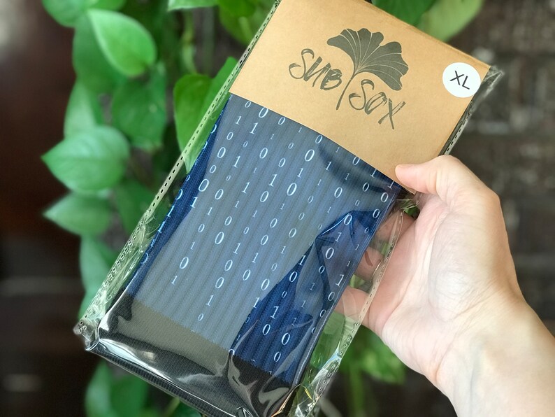 K&ouml;nnte beinhalten: Ein Paar blaue Socken mit einem wei&szlig;en Bin&auml;rcode-Muster. Die Socken sind in einer durchsichtigen Plastikt&uuml;te verpackt, die mit einem braunen Papieretikett mit der Aufschrift "SUB SOX" und "XL" versehen ist.