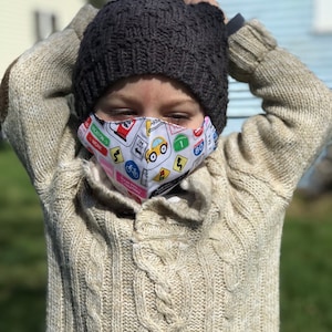 May include: A child wearing a beige cable knit sweater and a dark gray knit beanie. The child is wearing a face mask with a white background and colorful road signs and a yellow car.
