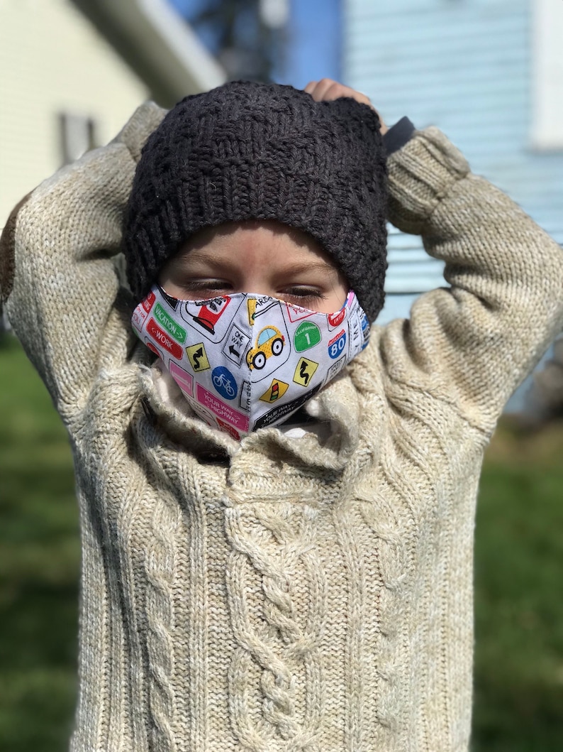 May include: A child wearing a beige cable knit sweater and a dark gray knit beanie. The child is wearing a face mask with a white background and colorful road signs and a yellow car.