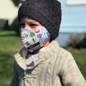 May include: A young child wearing a gray knitted beanie and a white face mask with a colorful road sign pattern. The mask features a yellow car, a stop sign, a yield sign, and a speed limit sign. The child is wearing a beige cable knit sweater.