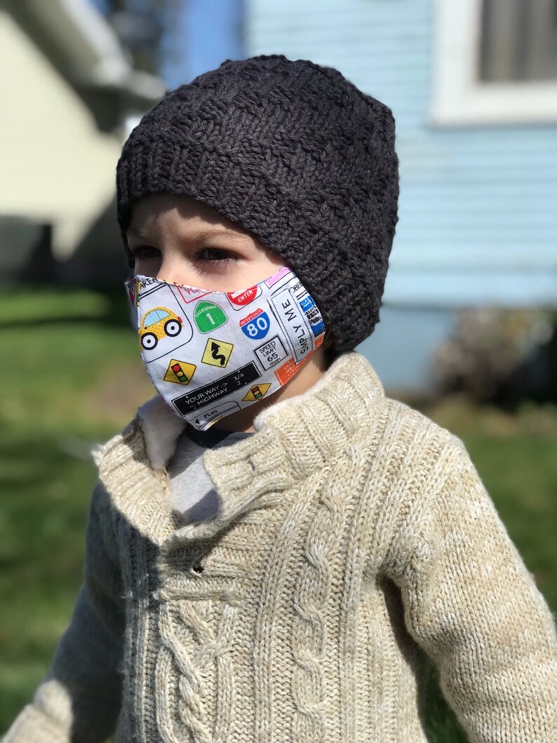 May include: A young child wearing a gray knitted beanie and a white face mask with a colorful road sign pattern. The mask features a yellow car, a stop sign, a yield sign, and a speed limit sign. The child is wearing a beige cable knit sweater.
