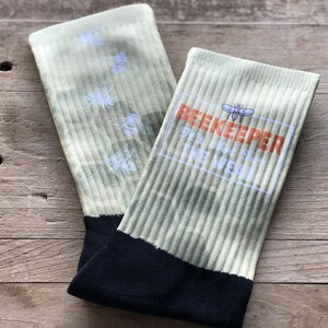 May include: A pair of green and white socks with a black heel and toe. The socks have a bee design and the text "BEEKEEPER OF THE VEIL" printed on them.