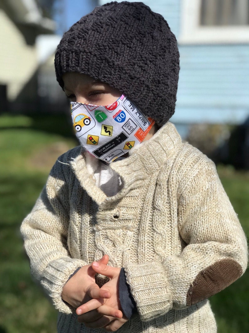 May include: A young child wearing a beige knitted sweater with brown elbow patches and a dark gray knitted hat. The child is wearing a face mask with a colorful pattern of traffic signs and the words "Simply Me" printed on it.