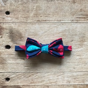 May include: Navy blue bow tie with a repeating red shark pattern. A turquoise fish is centered on the bow, with a turquoise band in the middle. The bow tie is set against a wooden background.