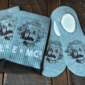 May include: A pair of blue socks with a white background featuring a repeated pattern of scientific equations and a black and white image of Albert Einstein. The socks have a black cuff and the text "E=MC2" is printed on the socks.