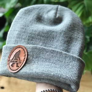 Op de afbeelding: Grijze gebreide beanie met een bruine leren patch met een silhouet van Bigfoot en de tekst "SPOKANE, WA".