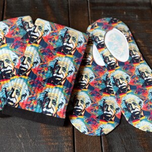 Könnte beinhalten: Socken und Einlegesohlen mit einem farbenfrohen Muster von Albert Einsteins Porträt. Die Socken haben einen schwarzen Bund, während die Einlegesohlen eine weiße obere Schicht haben. Das Design umfasst rote, blaue, gelbe und grüne Farbtöne.