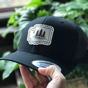 Puede incluir: Gorra de camionero negra con un parche plateado que presenta el contorno del estado de Washington con tres árboles de hoja perenne en su interior. El parche tiene un borde punteado. La gorra tiene una parte posterior de malla negra y una visera negra.