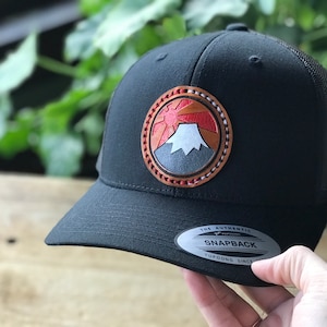 May include: Black trucker hat with a brown leather patch featuring a red sun rising over a gray mountain. The patch has white stitching around the edge. The hat has a black mesh back and a silver snap closure with the text "The Authentic Yupoong Snapback Yupoong Since" on it.