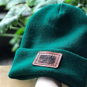 Pode incluir: Um gorro verde escuro com um remendo de couro marrom que apresenta um design de bandeira americana desbotado.
