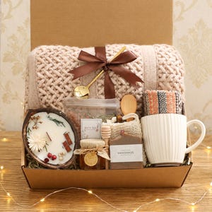 Könnte beinhalten: Eine Geschenkbox mit einer beigen Strickdecke, die mit einem braunen Band und einem goldenen Löffel verziert ist, einer Kokosnussschalen-Kerze, einem Glas Honig, Badesalz, einem Honiglöffel, einem Luffaschwamm, einem Stück Seife und einem weißen Becher.