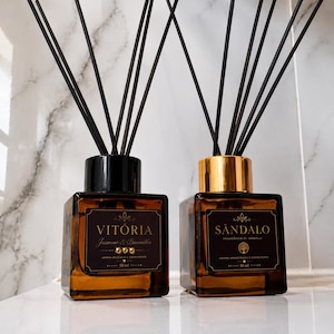 Pode incluir: Dois difusores de varetas em vidro âmbar quadrado com varetas pretas. O frasco esquerdo tem tampa preta e rótulo VITÓRIA Jasmin & Baunilha 50 ml. O frasco direito tem tampa dourada e rótulo SÂNDALO FRAGRÂNCIA DE SÂNDALO 50 ml.