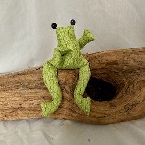 Könnte beinhalten: Ein Stofffrosch aus hellgrünem Stoff mit einem weißen Gänseblümchenmuster. Der Frosch hat zwei kleine schwarze Perlenaugen auf dem Kopf, lange baumelnde Beine und ausgestreckte Arme in einer sitzenden, aufrechten Position.