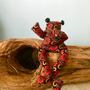 Könnte beinhalten: Eine handgefertigte Stofffroschpuppe mit einem Blumenmuster in Rot, Schwarz und Gold. Der Frosch hat schwarze Knopfaugen und ist auf einem Stück Treibholz platziert. Die Arme und Beine der Puppe sind ausgestreckt.