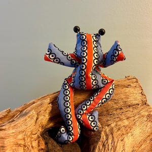 Könnte beinhalten: Ein Stofffrosch-Spielzeug mit hellblauem Grundton, einem wiederkehrenden Muster aus schwarzen Kreisen und einem markanten vertikalen orangefarbenen Streifen in der Körpermitte. Der Frosch hat zwei kleine schwarze Perlenaugen auf dem Kopf.