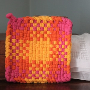 May include: A square, woven hot pad in shades of orange, yellow, and pink. The hot pad has a loop for hanging and is next to a stack of white fabric. The woven design creates a checkerboard pattern.