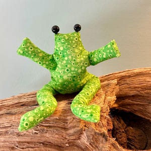 Könnte beinhalten: Ein Stofffrosch aus grünem Stoff mit einem Muster aus hell- und dunkelgrünen Kreisen. Der Frosch hat zwei schwarze Perlenaugen, ausgestreckte Arme und zwei ausgestreckte Beine. Der Stoff hat eine strukturierte Oberfläche mit sichtbaren Nähten.