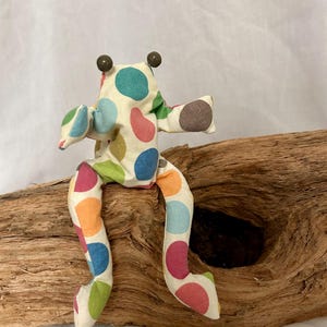 Könnte beinhalten: Ein Stofffrosch aus gepolstertem Material mit cremefarbener Basis und einem bunten Punktemuster in Türkis, Rosa, Orange, Blau und Grün. Der Frosch hat zwei kleine, dunkelbraune, kugelförmige Augen, die oben auf dem Kopf angebracht sind.