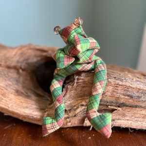 May include: A whimsical fabric frog doll with a plaid pattern in green, red, and tan. The frog has clear, round eyes and long, flexible legs. The doll is sitting on a piece of driftwood.