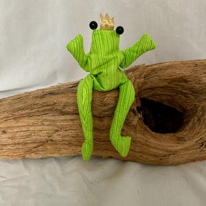 Könnte beinhalten: Ein Stofffrosch aus hellgrünem Stoff mit dünnen weißen vertikalen Streifen. Der Frosch hat zwei schwarze Perlenaugen, lange, herabhängende Arme und Beine sowie eine kleine goldfarbene Stoffkrone, die oben auf dem Kopf sitzt.