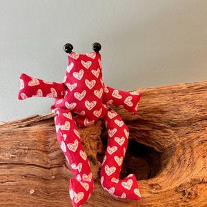 Könnte beinhalten: Ein Stofffrosch aus rotem Material mit einem sich wiederholenden weißen Herzmuster. Der Frosch hat zwei kleine schwarze, kugelförmige Augen auf dem Kopf, seitlich ausgestreckte Arme und lange Beine, die nach unten hängen.