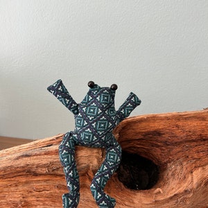 Könnte beinhalten: Eine handgefertigte Stoff-Frosch-Puppe mit einem geometrischen Muster in Teal, Marineblau und Weiß. Der Frosch hat dunkelbraune Perlenaugen und ist mit ausgestreckten Armen und Beinen posiert. Er sitzt auf einem Stück Treibholz.