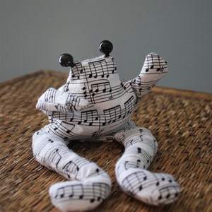 Könnte beinhalten: Ein weißes und schwarzes Stofffrosch-Plüschtier mit Notenaufdruck. Der Frosch hat schwarze, runde Augen und ist mit ausgestreckten Armen und Beinen posiert. Das Plüschtier sitzt auf einer gewebten, braunen Oberfläche.