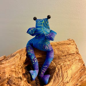Könnte beinhalten: Ein ausgestopftes Stofffrosch-Spielzeug mit einem leuchtend blauen und violetten Batikmuster mit grünen blattartigen Adern. Der Frosch hat zwei kleine schwarze Perlenaugen auf dem Kopf, lange baumelnde Beine und zwei nach außen gestreckte Arme.