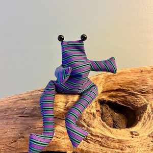 Könnte beinhalten: Ein Stofffrosch aus Textil mit horizontalen violetten und türkisfarbenen Streifen. Der Frosch hat zwei runde schwarze Perlenaugen auf dem Kopf, lange Gliedmaßen und eine sitzende Haltung, bei der ein Arm seitlich nach außen gestreckt ist.